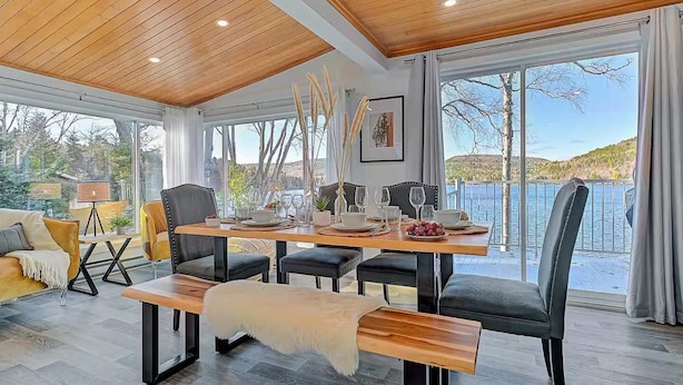 Un comedor con una mesa de madera dispuesta para cuatro personas, rodeado de sillas, un banco y vistas a un lago.
