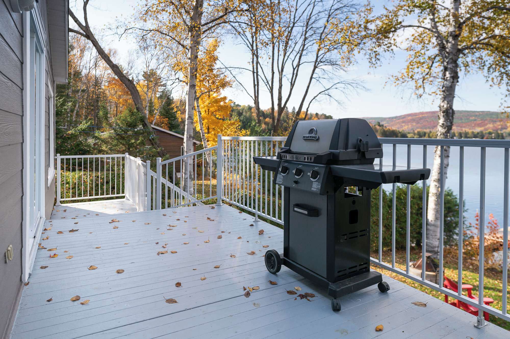 Une terrasse dotée d’un barbecue mobile, d’une rambarde blanche et offrant une vue sur un lac et les arbres aux couleurs automnales.