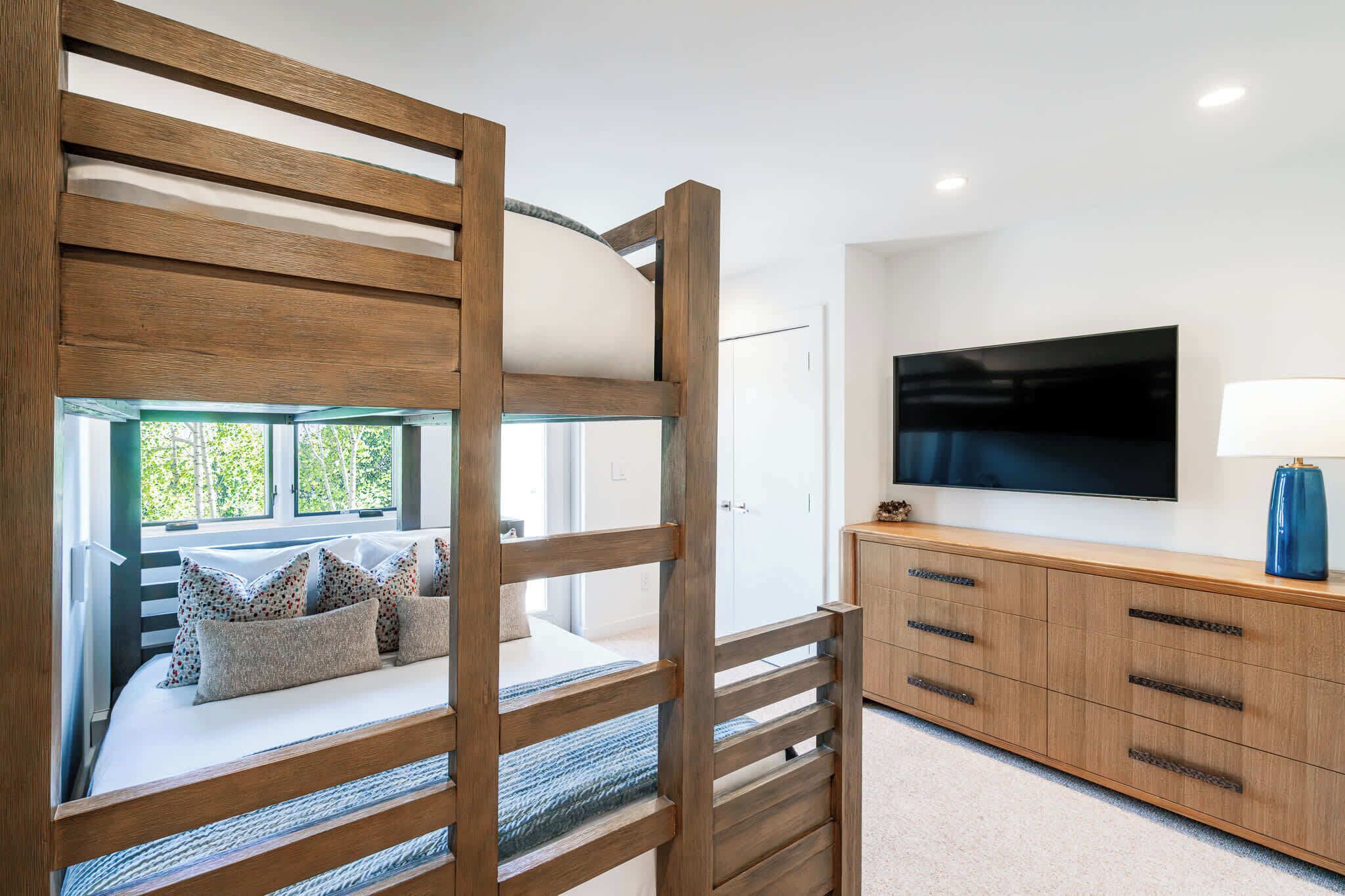 A bunk bed with a view of greenery, a TV on the wall, and a wooden cabinet with drawers.