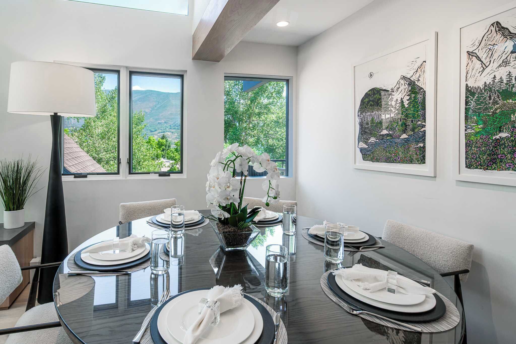 A dining area with a round table set for four, surrounded by chairs, with a view of greenery through large windows.