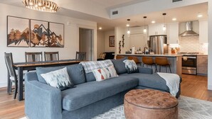 A modern living room with a sectional sofa, ottoman, dining area, kitchen, and artwork on the wall. - TimberFox 3209 | 2BR Downtown, Hot Tub, A/C (Winter Park)