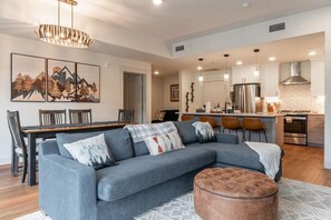 A modern living room with a sectional sofa, ottoman, dining area, kitchen, and artwork on the wall.