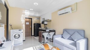 A modern living room with a grey sofa, a washing machine, and a kitchen area with a dining table and chairs. - Condo Gym Plane Views Near Beach C172 (Mai Khao)