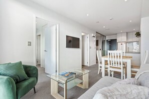 A modern living room with a glass coffee table, a green sofa, and a white sofa. There is a dining area with a wooden table and white chairs. The kitchen is visible with white cabinets and a refrigerator.