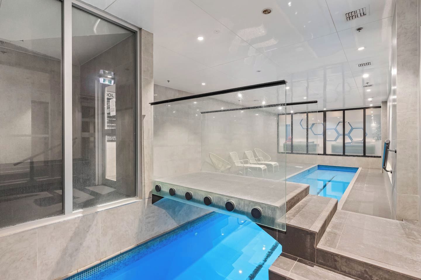 A modern indoor swimming pool with clear glass walls, a diving board, and lounge chairs.