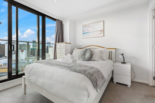 A hotel room with a large window, a bed with white bedding, a bedside table with a lamp, and a city view.