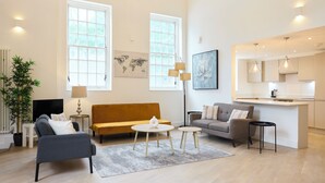A modern living room with a sofa, two chairs, a coffee table, and a small side table. There is a large window, a world map on the wall, and a kitchen area visible through an open door. - Central Tadworth Flat (Tadworth)