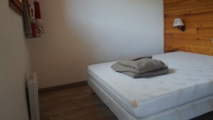 Quarto pequeno com uma cama de solteiro, cabeceira de madeira, colchão branco e uma mesinha de cabeceira com abajur.