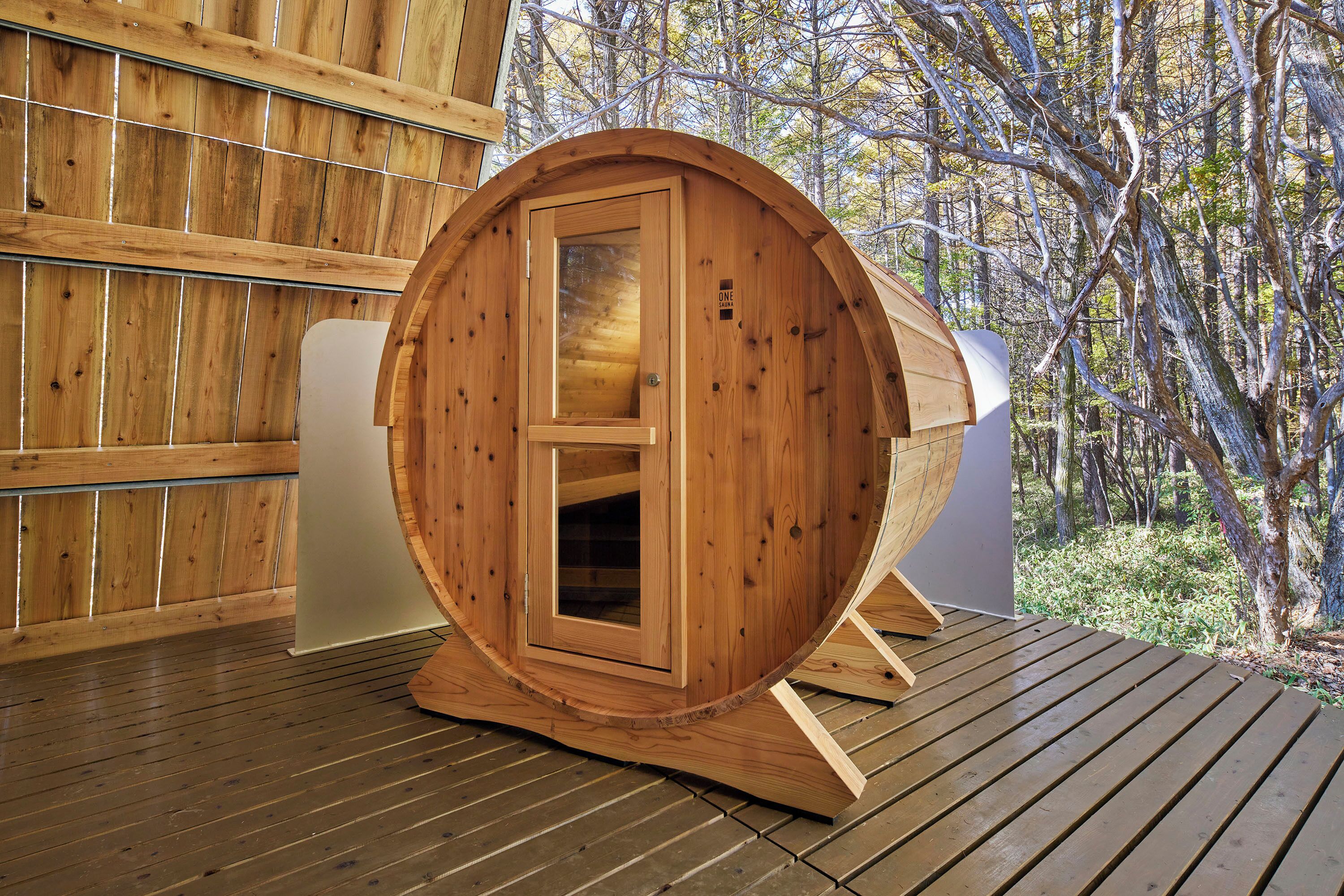 A wooden sauna with a door, set on a wooden deck with a forest backdrop.
