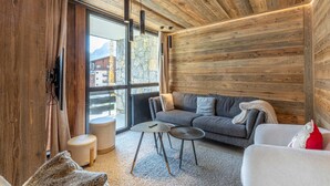 Ein modernes Interieur mit Holzboden, einem grauen Sofa, einem runden Tisch und einem großen Fenster mit Blick auf Gebäude.