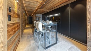 Ein schmaler Flur mit Holzwänden und -decke, der zu einem Essbereich führt, mit einem Tisch für vier Personen, Metallstühlen und einer schwarzen Arbeitsplatte mit integriertem Backofen.