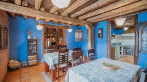 Un coin repas rustique avec des poutres en bois, un mur bleu et une cuisine visible à travers une porte ouverte.