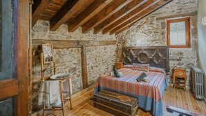 Une chambre à coucher rustique avec un mur en pierre, des poutres en bois, un lit avec une literie à motifs, une petite table et une valise.