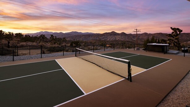 An outdoor tennis court with a net, surrounded by a fence, and set against a mountainous backdrop during sunset. - Hoopa House by Fieldtrip - Pickleball + 2 Pools (Yucca Valley)