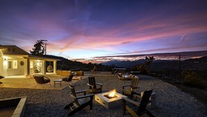 An outdoor seating area with fire pit, chairs, and tables, overlooking a scenic landscape with mountains and a sunset. - Hoopa House by Fieldtrip - Pickleball + 2 Pools (Yucca Valley)