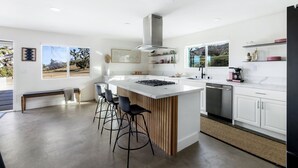 A modern kitchen with a central island, bar stools, a dishwasher, and a large window offering a view of the outdoors. - Hoopa House by Fieldtrip - Pickleball + 2 Pools (Yucca Valley)