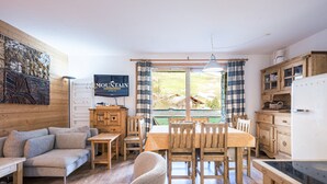 En hyggelig stue med sofa, spisebord med stole, et tv på væggen og et køkken med træskabe og et køleskab.