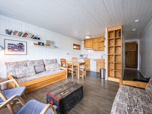Uma sala de estar acolhedora com sofá, cadeiras, uma mesa de madeira e uma cozinha ao fundo.
