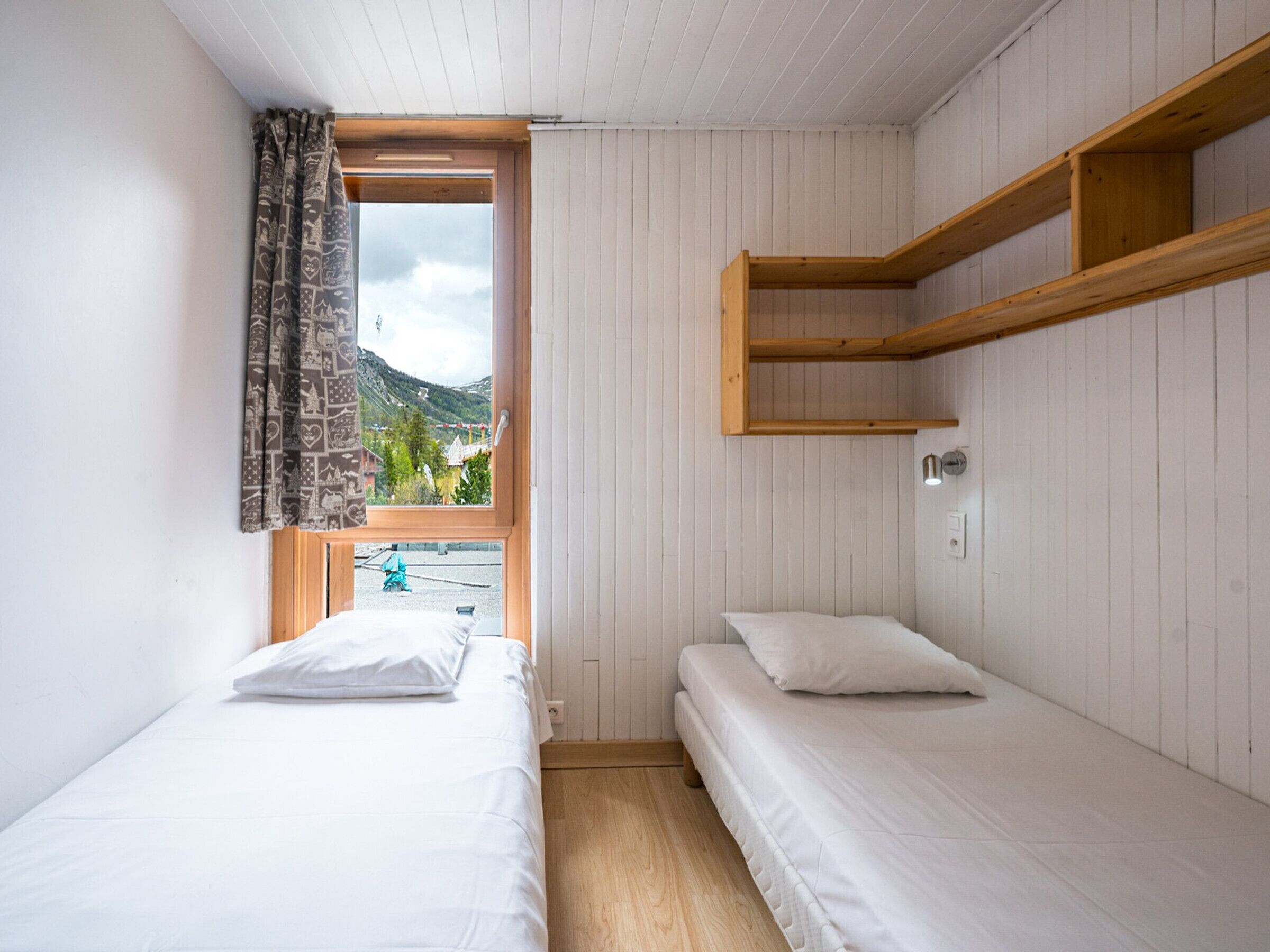 A room with two beds, a window with curtains, a wooden shelf, and a view of a mountain.