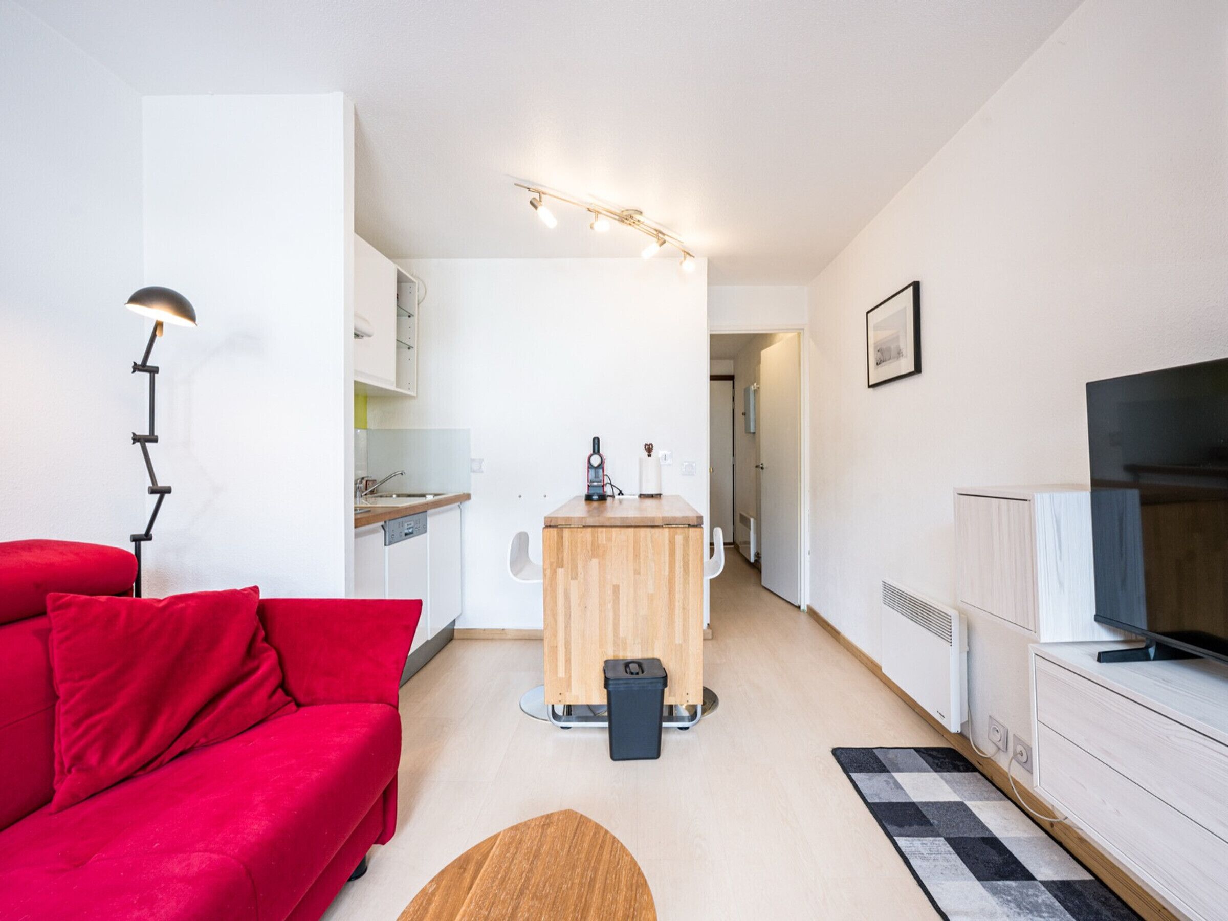 A compact living space with a red sofa, a small wooden table, a kitchenette, and a TV.