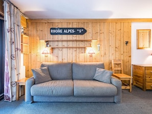 Um quarto aconchegante com um sofá cinza, paredes de madeira, um letreiro com a inscrição 'RHONE ALPES - 'S', duas luminárias fixadas na parede e uma pequena mesa de madeira.