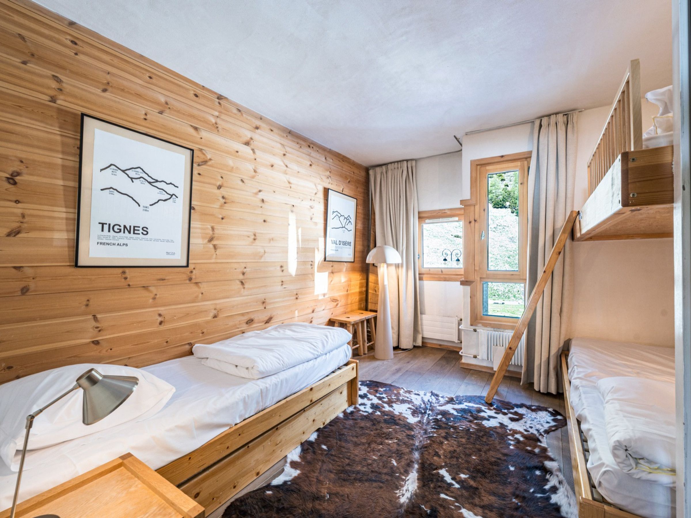 A room with wooden walls, a bed, a desk, a lamp, a cowhide rug, and a window with curtains.
