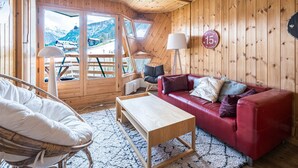 Een gezellige kamer met een rode bank, een houten salontafel en een rieten stoel. Een groot raam biedt uitzicht op bergen.