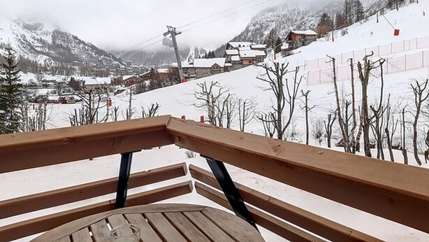 Een houten bank met uitzicht op een besneeuwd bergdorpsgezicht, met huizen en een skilift.