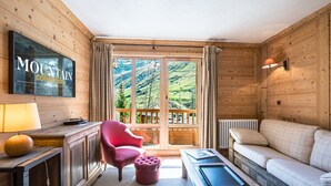A cozy room with a wooden interior, a television mounted on the wall, a sofa, a pink chair, a small table, and a view of a mountainous landscape through the window. - Charming Apartment in Chatelard, Val-d'Isère, Direct Access to Slopes, Parking Included (Val-d'Isère)