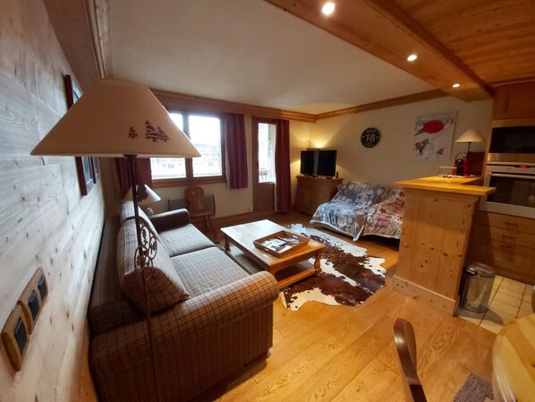 A cozy living room with a sofa, a coffee table, and a cowhide rug. There is a kitchen area with a stove and cabinets. - Cozy apartment with direct slope access, parking, and Bellevarde view (Val-d'Isère)