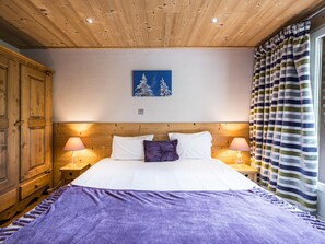 A bedroom with a wooden ceiling, a bed with white and purple bedding, two bedside lamps, a wooden wardrobe, and a window with striped curtains. - Cozy renovated apartment for 2-4 guests at ski slopes Val-d'Isère (Val-d'Isère)