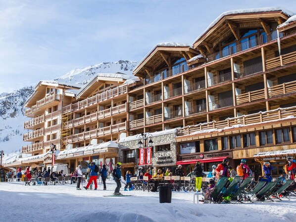 A multi-story wooden building with balconies, ski equipment, and a snow-covered mountain in the background.