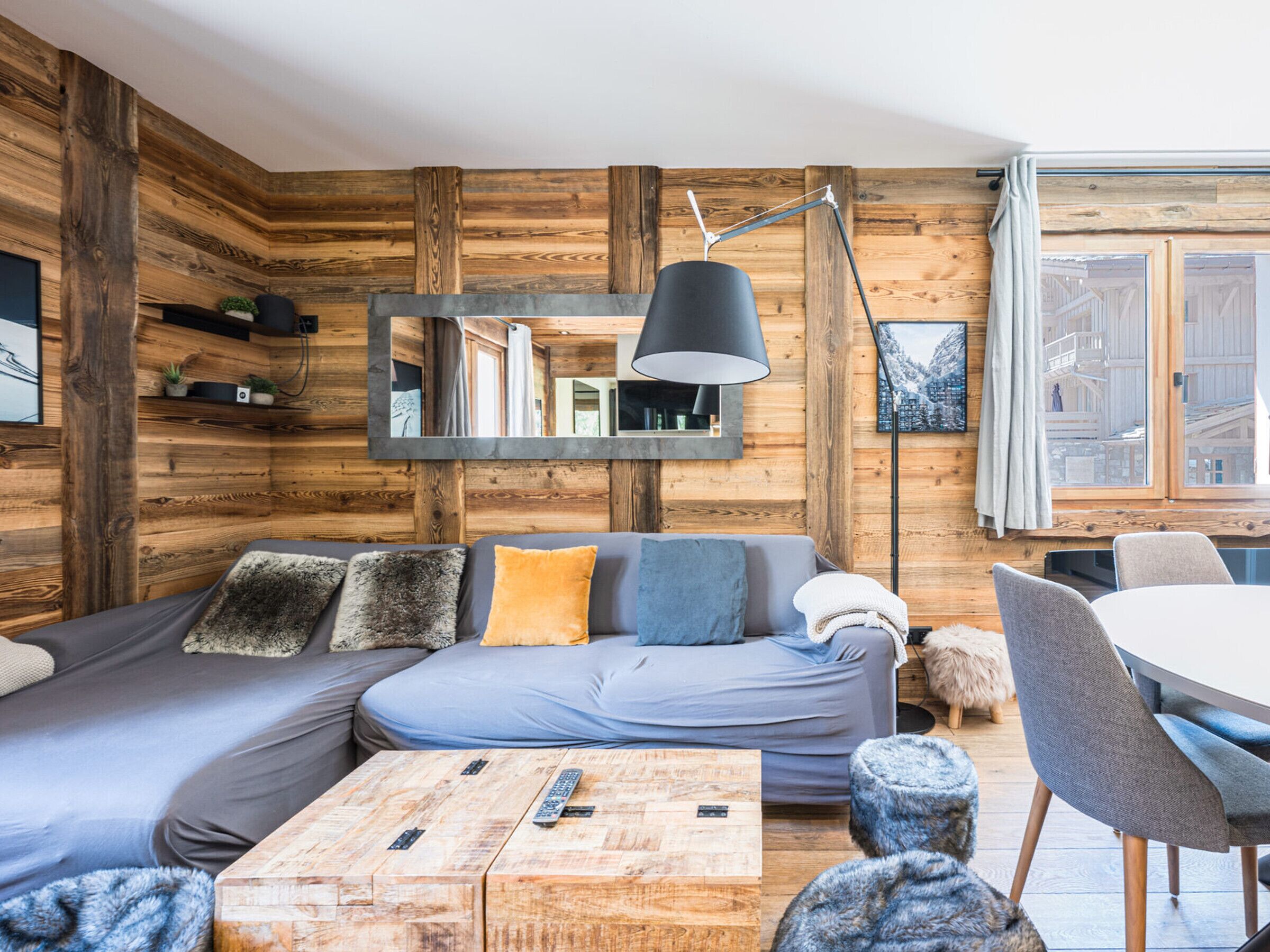 A modern living room with a wooden wall panel, a sofa with cushions, a coffee table with remotes, and a dining area with chairs.