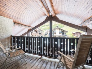 Una terraza de madera con dos sillas, una barandilla con detalles decorativos y vistas a edificios al fondo.