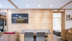 A modern interior with a wooden wall, a dining table with chairs, a bed, and a mounted sign. - Luxury 3-room apartment, central location, parking included (Val-d'Isère)