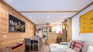 A modern interior with wooden walls, a dining area, a kitchen, and a living room with a sofa and a TV mounted on the wall. - Luxury 3-room apartment, central location, parking included (Val-d'Isère)