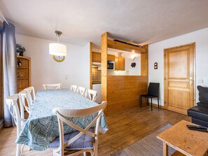 A dining area with a table and chairs, a kitchen with wooden cabinets, a black chair, a wooden door, and a small table with a remote control.