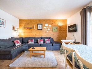 A cozy living room with a wooden wall, a grey sofa, a wooden coffee table, and a TV on the wall.