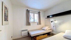 Een kleine kamer met een eenpersoonsbed, een bureau, een raam met gordijnen, een radiator en een wandplank.