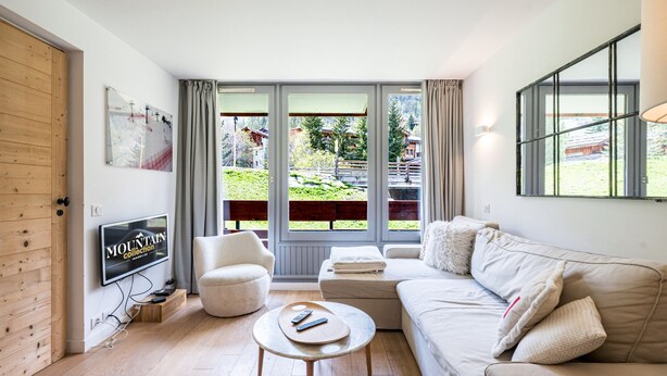A modern living room with a sofa, ottoman, and coffee table, a TV, and large windows with a view of greenery.
