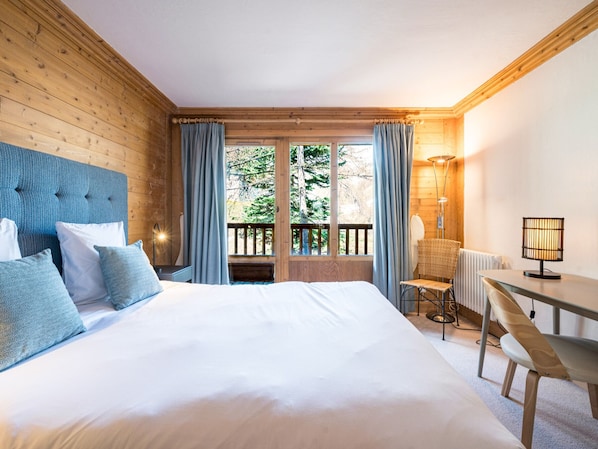 A bedroom with a large bed, a chair, a table, a lamp, and a view of trees through a window.