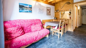 A cozy room with a wooden ceiling, a pink sofa with a patterned cover, a dining table with chairs, and a small kitchen area. - Cozy mezzanine studio in Val-d'Isère center (Val-d'Isère)