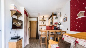A cozy room with a wooden dresser, a television, a dining table with chairs, a bed with patterned bedding, and a kitchen area with red tiles. - Renovated Studio Cabin - Ski-in/Ski-out - WIFI (Val-d'Isère)