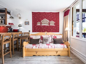 A cozy room with a wooden sofa, a dining table with chairs, a red tapestry with a house design, and a window with curtains.