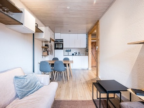 A compact living space with a sofa, a dining table with chairs, a kitchenette, and a wooden ceiling.