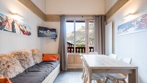 A modern room with a sofa, dining table, and chairs, a TV mounted on the wall, and a view of the outdoors through a window.