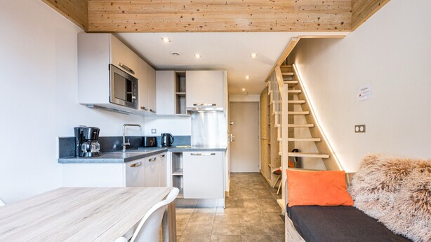 A compact kitchen with modern appliances, a dining area with a wooden table, and a staircase leading to a loft bedroom.