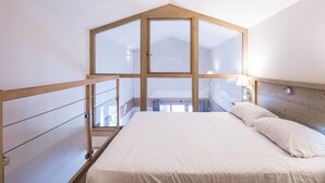 A bedroom with a sloped ceiling, a bed with white linens, a wooden headboard, and a window with a view of the outside.