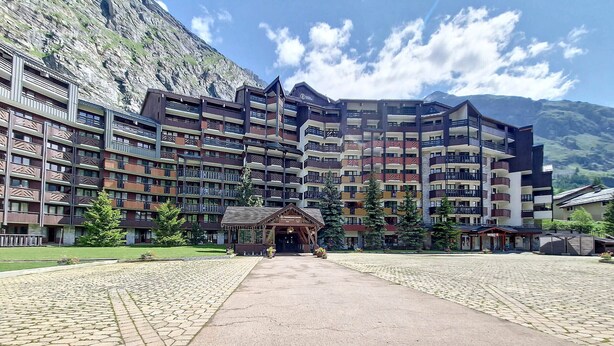 A multi-story hotel with a central entrance, surrounded by a cobblestone courtyard and a mountainous backdrop.