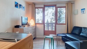 A room with a wooden dresser, a TV, a small table with a lamp, a blue sofa, and a view of a snowy landscape through a glass door.